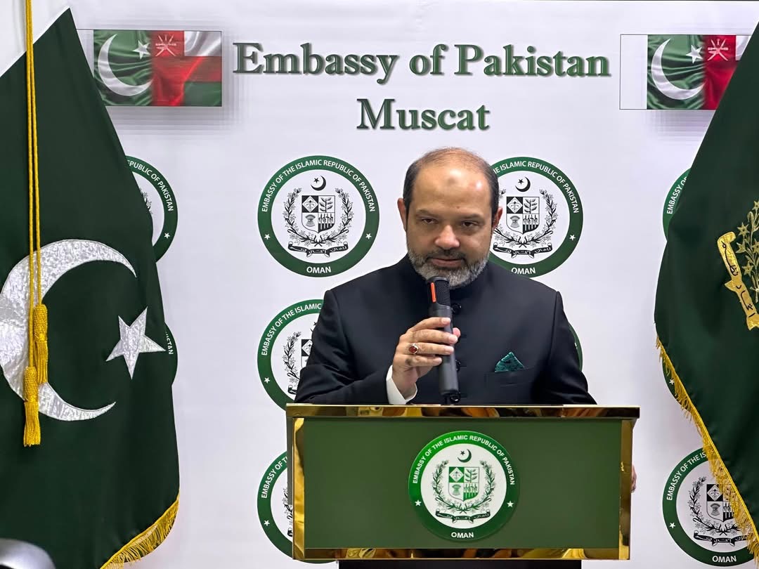 Ambassador of Pakistan to Oman Syed Naveed Safdar Bokhari addressing the 80th Independence Day ceremony at the Embassy in Muscat, flanked by Pakistan flags.